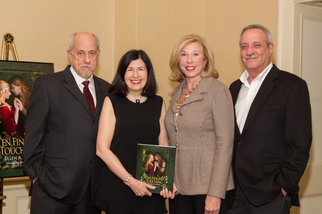 Group of four adults standing indoors, smiling. One holds a book titled "Ten Fingers Touching." A poster of the book is displayed behind them. Professional setting.