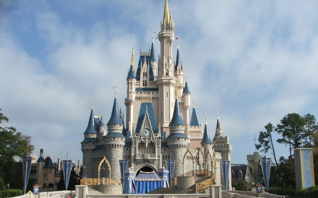 A majestic fairy tale castle with blue and gold spires towers against a cloudy sky. Elegant arches and flags add a magical ambiance to the scene.