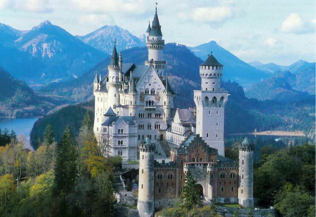 Neuschwanstein Castle towers over a lush forest, set against the backdrop of rugged blue mountains and a bright sky, evoking a fairy-tale allure.