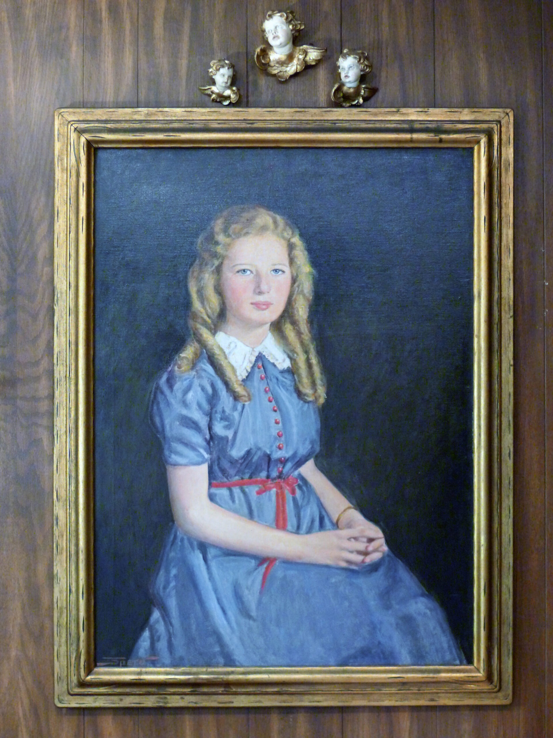 Portrait of a young girl with blond hair wearing a blue dress with red buttons and ribbon. The frame is ornate, with cherub sculptures above, on a wooden background.