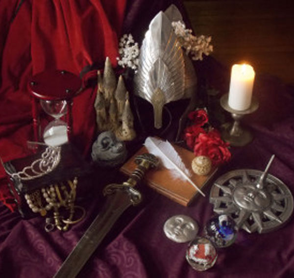 A mystical still life scene featuring a silver helmet, a lit candle, an hourglass, a quill on a notebook, a sword, and red roses on rich fabric.