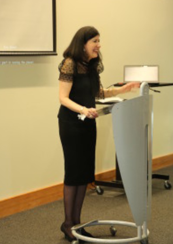 A woman in a black dress speaks passionately at a podium in a conference room. She appears engaged, with a projector in the background and a serious expression.